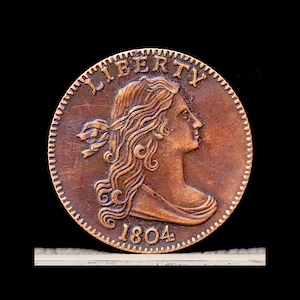 May include: A copper-colored coin with the word "LIBERTY" arched across the top. The coin features a profile of a woman with flowing hair and the date "1804" at the bottom. The coin has a textured edge and is set against a black background.