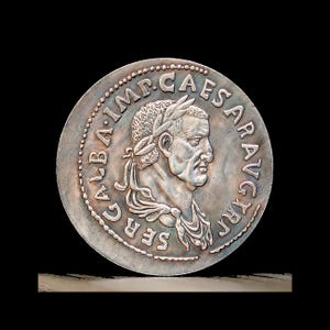 May include: A circular silver-toned coin featuring a profile relief of a person wearing a laurel wreath. The text SER GALBA IMP CAESAR AVG TR P is embossed around the perimeter, accompanied by a beaded border design on the coin's edge.