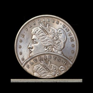 May include: A silver coin with a detailed profile of a woman, surrounded by the inscriptions "E PLURIBUS UNUM" and "LIBERTY". The coin has a textured surface and is set against a black background. The coin's design includes stars and floral elements.