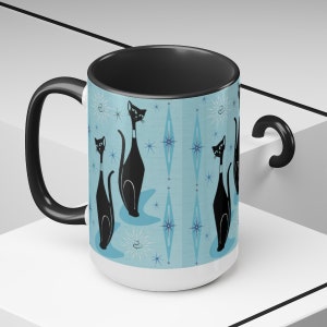 May include: A white ceramic mug with a black handle and a black rim. The mug features a repeating pattern of black cats with blue eyes and a blue and white geometric design.