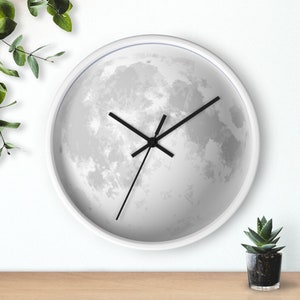May include: A round white wall clock featuring a detailed image of the moon. The clock has black hands and a white frame. The clock is displayed on a white wall with a small potted plant and greenery.