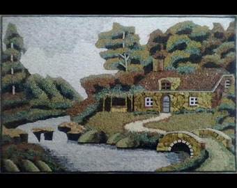 Puede incluir: Una obra de arte de mosaico de piedra que representa una cabaña pintoresca enclavada en un exuberante bosque verde. Un camino sinuoso conduce a un puente de piedra sobre un arroyo. La obra está enmarcada y tiene un encanto rústico.