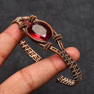 May include: A copper-coloured wire-wrapped bracelet with a large, oval-shaped, faceted red gemstone. The bracelet has a woven wire design on the band. The gemstone is the focal point. The bracelet is held in a hand against a dark background.