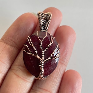 Könnte beinhalten: Ein roter Edelstein-Anhänger mit einem Baumdesign aus Kupferdraht. Der Anhänger ist oval geformt und hat eine Öse aus Kupferdraht.