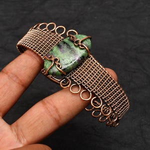 May include: Copper wire bracelet with a rectangular green and red stone. The bracelet has a woven design with circular accents. The bracelet is displayed against a dark background.