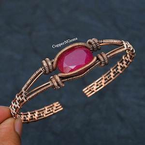 May include: A wire-wrapped cuff bracelet made of copper featuring a central oval-shaped faceted pink gemstone. The metalwork consists of intricate coiled and woven wire patterns. The text CopperNGems is visible above the central stone.
