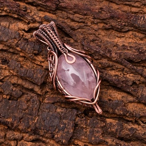 Könnte beinhalten: Ein Anhänger aus rosa Rosenquarz, der in Kupferdraht gewickelt ist. Der Anhänger ist ein zartes und filigranes Design mit einem natürlichen, organischen Look.