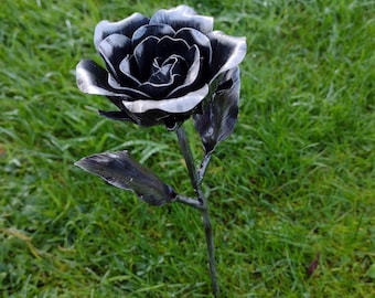 May include: A handcrafted metal rose sculpture with a dark gray finish. The rose has detailed petals and leaves, and is set against a backdrop of green grass. The sculpture is a decorative garden accent.