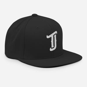 May include: Black baseball cap with a flat peak, featuring a white embroidered letter "J". The cap has a structured crown. The design is simple and modern, suitable for casual wear. The cap is a versatile accessory.