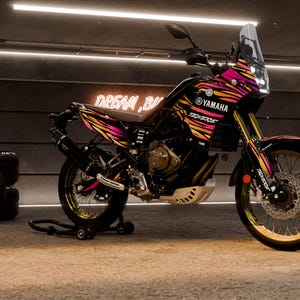 May include: A black Yamaha motorcycle with a vibrant pink, orange, and yellow striped design. The motorcycle has gold-colored accents on the wheels and suspension. The word "Yamaha" is visible on the side.