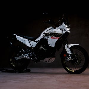 May include: A white and black Yamaha Ténéré 700 motorcycle with a distinctive graphic design. The motorcycle features the word "Ténéré" in bold lettering, along with the Yamaha logo. The wheels have a gold rim. The motorcycle is parked on a stand.