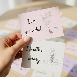 Könnte beinhalten: Zwei Karten mit inspirierenden Botschaften werden gehalten. Die obere Karte ist rosa mit den Worten "I am grounded" und einer Strichzeichnung einer Frau mit Blumen. Die untere Karte ist weiß mit "I breathe calmly" und einer meditierenden Frau.