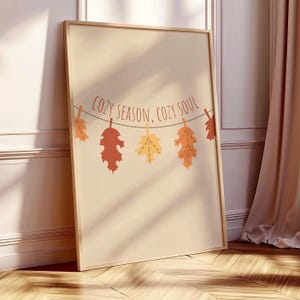 Può includere: Una stampa artistica incorniciata con sfondo beige chiaro che presenta una ghirlanda di cinque foglie autunnali appese nei toni dell'arancione, del rosso e del giallo. Il testo "COZY SEASON, COZY SOUL" è stampato in marrone sopra la ghirlanda.
