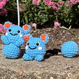 May include: Two blue crochet figures with orange ears and a blue crochet ball sit on a stone surface. The figures have white accents and smiling faces. Pink flowers and greenery are in the background.