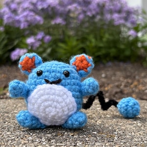 May include: A crocheted blue creature toy with orange ear details and a white belly. It has a black tail and a blue ball at the end. The toy is placed on a concrete surface, with purple flowers visible in the background. A handmade item.