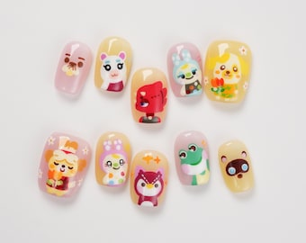 May include: A set of ten press-on nail tips featuring colorful cartoon character illustrations. The designs include various animals and creatures in shades of yellow, pink, green, and red, with some tips decorated with small white flower accents.