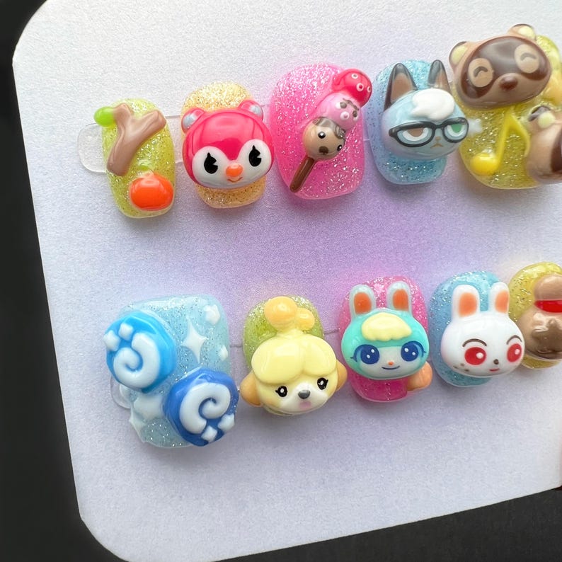 May include: A set of ten artificial nails with various 3D character designs. The nails feature a variety of colors, including pink, yellow, blue, and white, with glitter accents. The designs include cartoon characters and food items.