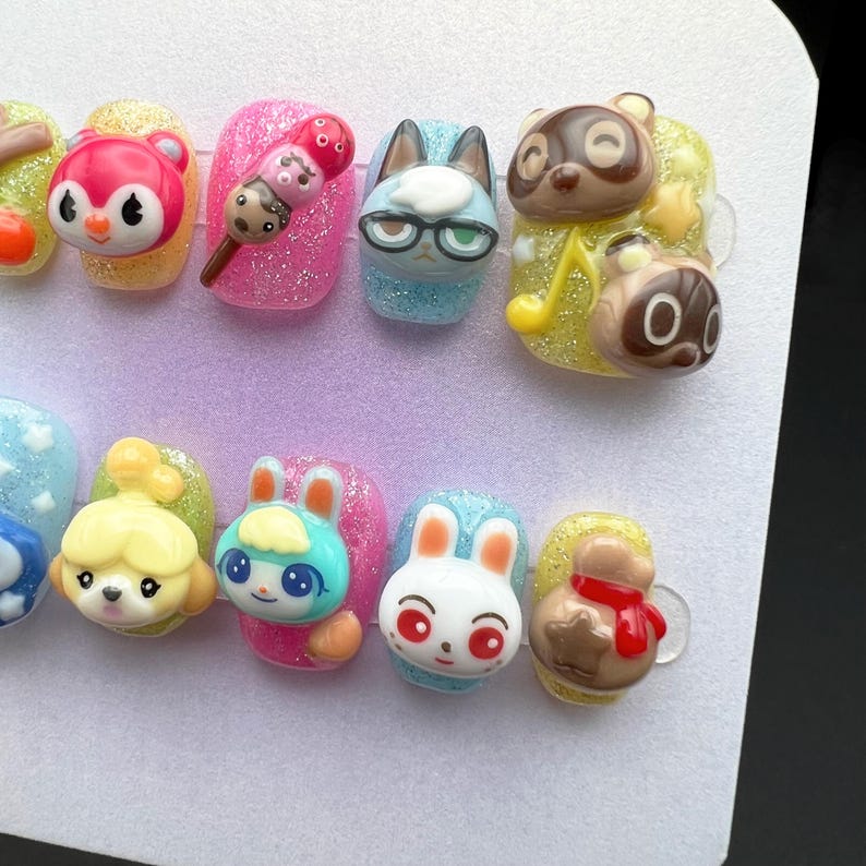 May include: A set of custom-designed press-on nails featuring various cartoon character designs. The nails showcase a variety of colors, including pink, blue, yellow, and white, with glitter accents. Each nail has a unique 3D character design.