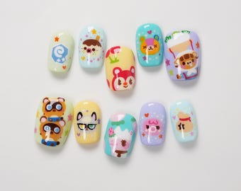 May include: A set of ten press-on nails featuring colorful designs inspired by Animal Crossing characters. Each nail has a glossy finish with various illustrations including tanuki, cats, bears, and game-related icons in pastel shades.