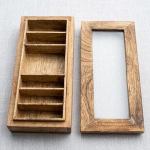 Op de afbeelding: Een rechthoekige houten doos met een deksel, met een reeks kleine compartimenten. Het deksel is een houten frame met een transparante inzet. Het hout heeft een natuurlijke, lichtbruine kleur en zichtbare nerf.
