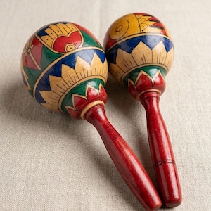 May include: A pair of wooden maracas with red handles and decorated spherical heads. The heads are painted with red, green, blue, and yellow patterns. The maracas are displayed on a neutral-coloured fabric surface.