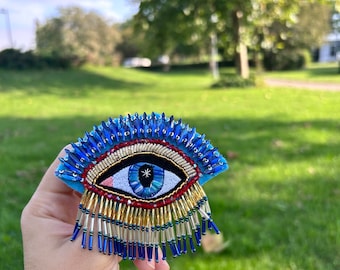 May include: A handmade, embroidered eye-shaped brooch. The brooch features a blue eye with a star-shaped pupil, surrounded by gold and red beadwork. Blue crystal-like beads fringe the bottom and top of the brooch. The background is a blurred outdoor scene.