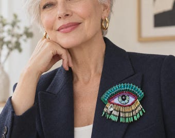 Puede incluir: Un broche de ojo bordado con un iris azul central, rodeado de capas de bordado en color verde azulado, rojo y negro. El diseño incluye flecos metálicos colgantes en tonos verde, dorado y plateado, prendido en un blazer azul marino oscuro.