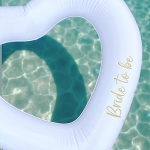 May include: White heart-shaped inflatable pool float with the text "Bride to be" in gold lettering.
