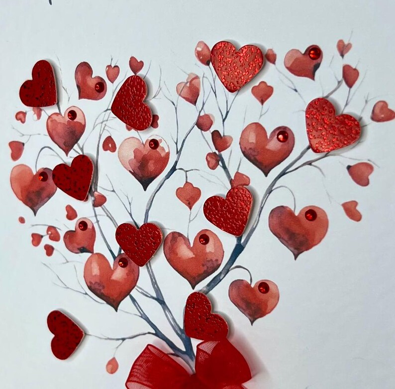 May include: A white card featuring a watercolour painting of a tree with red hearts. The tree has a red ribbon bow at its base. The hearts are painted in various shades of red and some have glitter.