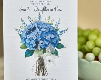 Op de afbeelding: Een witte trouwkaart met een illustratie van een blauw hortensia boeket. De kaart leest "WITH LOVE TO A VERY SPECIAL Son & Daughter in Law" en "ON YOUR WEDDING DAY Oscar & Olivia 20th JUNE 2025". Het boeket is vastgebonden met een wit lint.