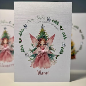 May include: A Christmas card with a watercolor illustration of a fairy in a pink dress and wings, surrounded by a wreath of greenery and stars. The card reads "Merry Christmas To A Wonderful Granddaughter" and has the name "Niamh" at the bottom.