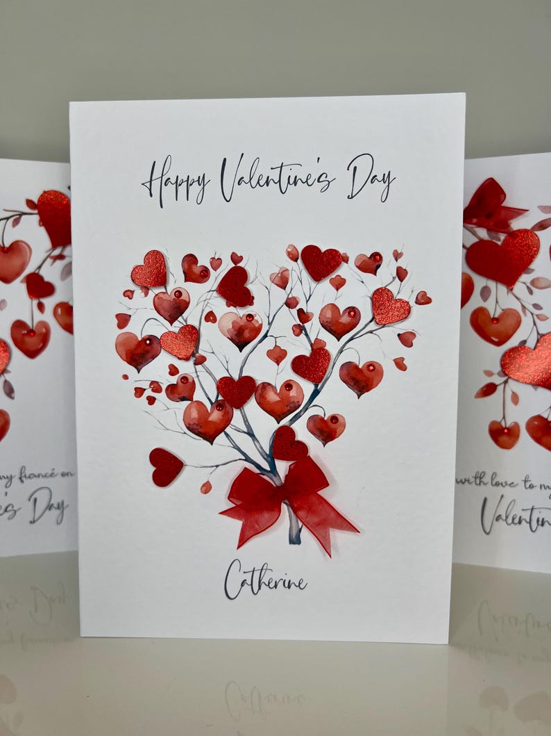 May include: A white card with the text "Happy Valentine's Day" and a red heart-shaped tree with a red bow. The name "Catherine" is written below the tree.