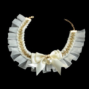 May include: A white and gold necklace with a pearl and bow accent. The necklace is made of a delicate fabric with a ruffled edge.