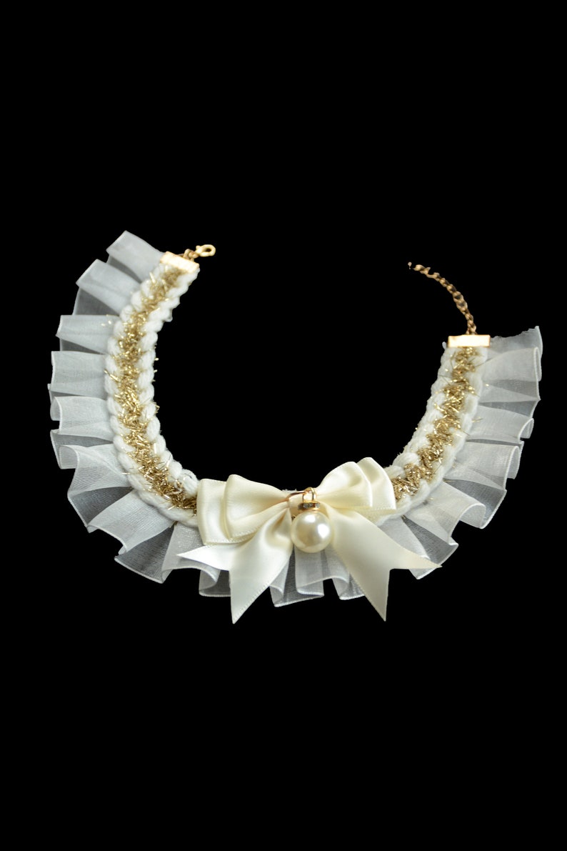 May include: A white and gold necklace with a pearl and bow accent. The necklace is made of a delicate fabric with a ruffled edge.