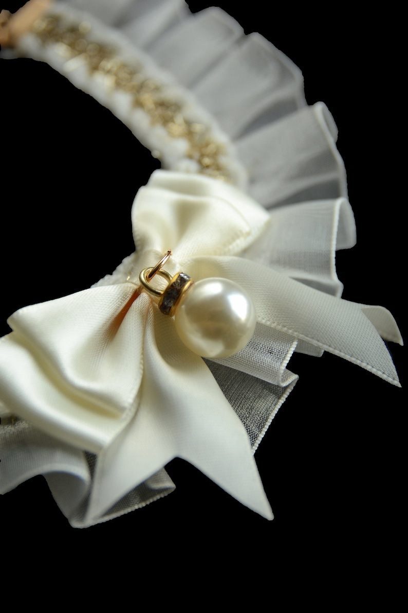 May include: A white satin bow with a pearl and rhinestone embellishment, attached to a white tulle collar.