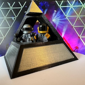 May include: A black and gold pyramid-shaped sculpture featuring two miniature figures with helmets and microphones. The figures are dressed in black and white patterned suits. The sculpture has a silver and gold metallic finish.