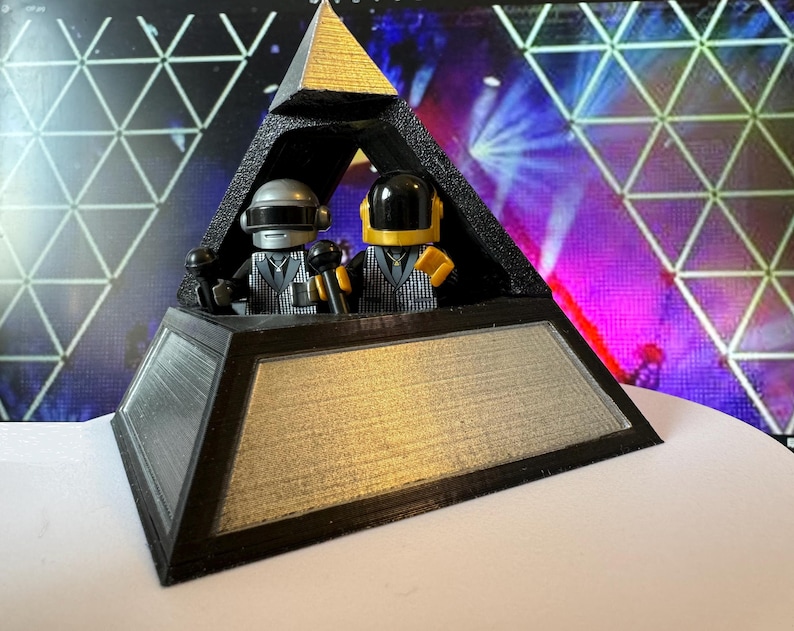 May include: A black and gold pyramid-shaped sculpture featuring two miniature figures with helmets and microphones. The figures are dressed in black and white patterned suits. The sculpture has a silver and gold metallic finish.