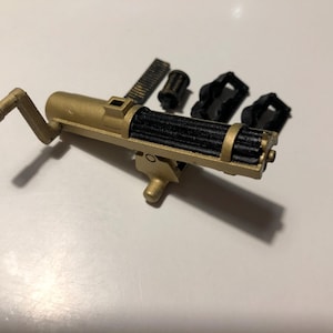 Puede incluir: Una ametralladora de juguete de color dorado con un ca&ntilde;&oacute;n negro y una manivela. La pistola est&aacute; desmontada e incluye varias piezas adicionales.