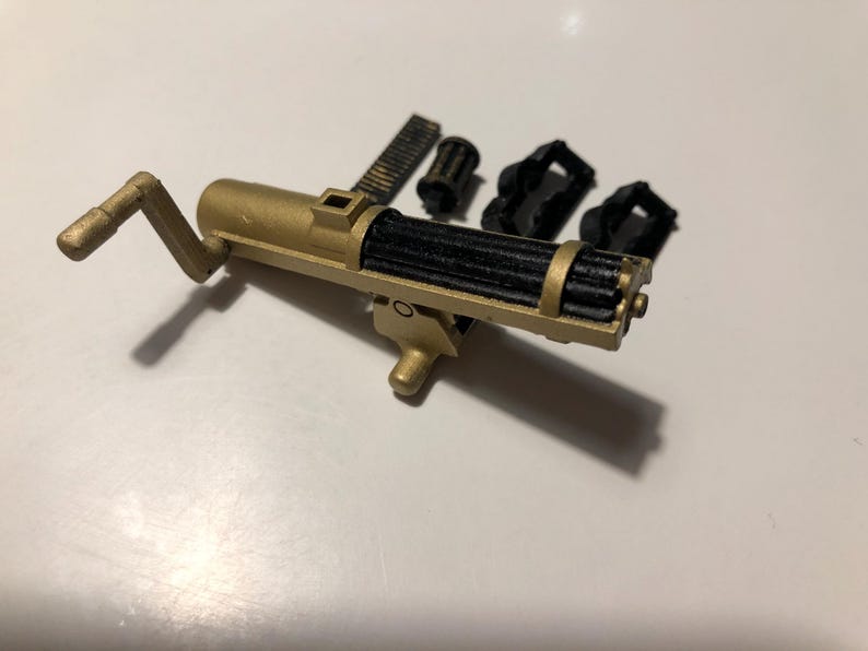Puede incluir: Una ametralladora de juguete de color dorado con un ca&ntilde;&oacute;n negro y una manivela. La pistola est&aacute; desmontada e incluye varias piezas adicionales.
