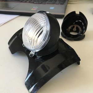 May include: A black motorcycle headlight with a clear lens and a black plastic housing. The headlight is mounted on a black metal bracket.