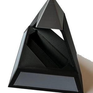 May include: A black and gray geometric pyramid-shaped organizer. The organizer has a triangular top and base with a central compartment. Gray accents are on the base and top. The design is modern and minimalist.