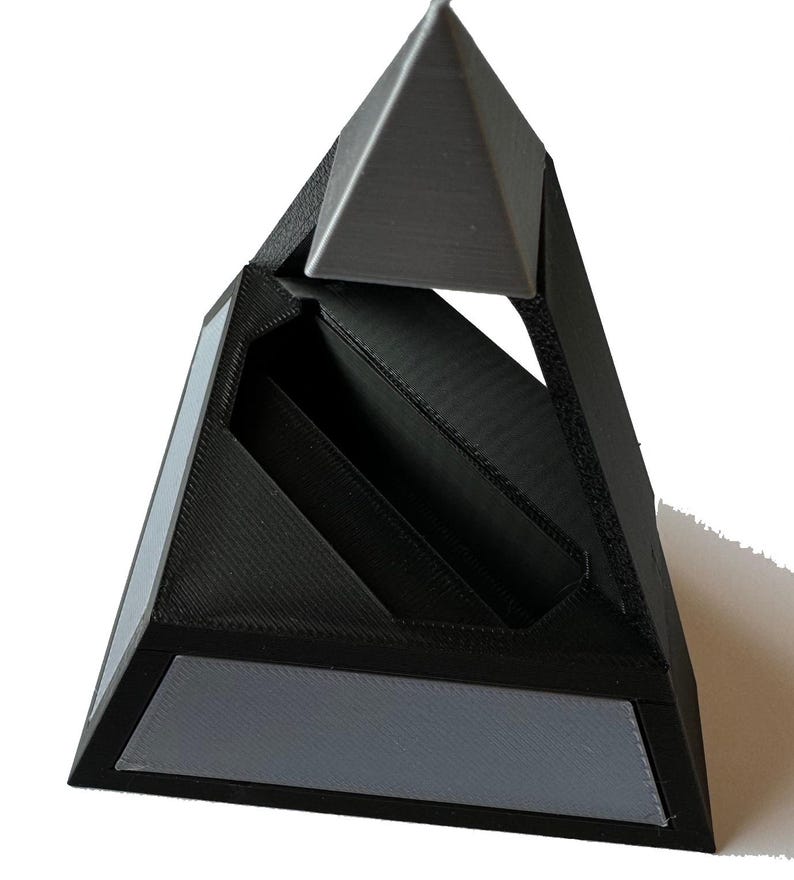 May include: A black and gray geometric pyramid-shaped organizer. The organizer has a triangular top and base with a central compartment. Gray accents are on the base and top. The design is modern and minimalist.