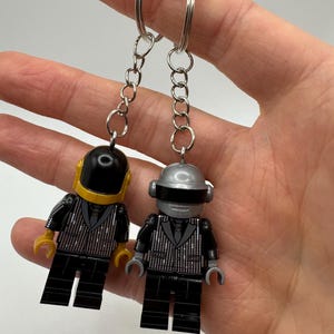 Könnte beinhalten: Zwei silberne und schwarze Lego-Minifiguren mit Helmen und Anzügen, die an silbernen Schlüsselanhängern befestigt sind. Die Minifiguren tragen schwarze Anzüge mit silbernen Akzenten und haben schwarze Beine.