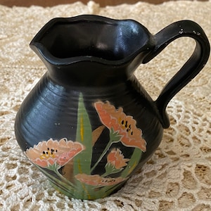 May include: A black ceramic pitcher with a floral design. The flowers are painted in shades of pink, orange, and yellow. The pitcher has a wide mouth and a handle.