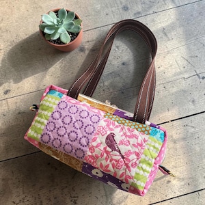 May include: A patchwork fabric handbag with a bird design, featuring pink, purple, green, and gold patterns. The bag has brown handles and a gold clasp. A small potted succulent is in the background.
