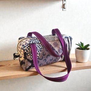 May include: A patchwork fabric tote bag with a purple strap and white stitching. The bag features a variety of patterns in shades of black, gray, and beige. The bag is sitting on a wooden shelf with a small potted plant.