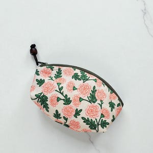 May include: A small, oval-shaped pouch with a floral pattern. The pouch features a cream-colored background with pink flowers and green leaves. It has a dark green zipper and a small, wooden bead on the zipper pull.