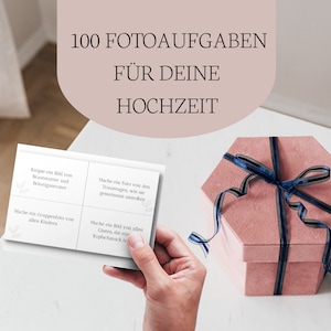 Puede incluir: Una caja rosa con una cinta azul atada alrededor. La caja está sentada en una mesa blanca. Hay dos tarjetas en la mesa con texto que dice "100 Fotoaufgaben für deine Hochzeit".