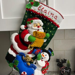 May include: A green felt Christmas stocking featuring a white cuff embroidered with the name Elaina. The stocking displays a Santa Claus holding a yellow list, a snowman with a black hat, a blue gift, a teddy bear, and a candy cane, all accented with sequins.