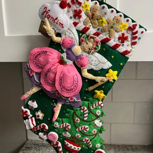 Könnte beinhalten: Ein handgefertigter Weihnachtsstrumpf mit grüner Filzbasis, verziert mit einer Fee, Lebkuchenmännern, Zuckerstangen und Cupcakes. Die Fee trägt ein rosa Kleid und hält einen Stern. Der Name "Hanlim" ist auf einer weißen Wolke gestickt.